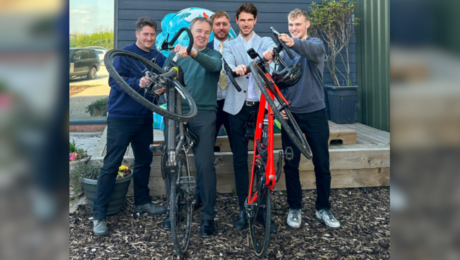 Group of gentleman holding 2 bikes, one black, one red, in front of decking