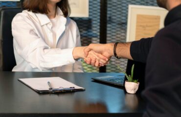 Two business leaders shaking hands across a table with a laptop and clipboard