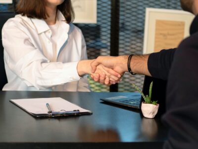 Two business leaders shaking hands across a table with a laptop and clipboard