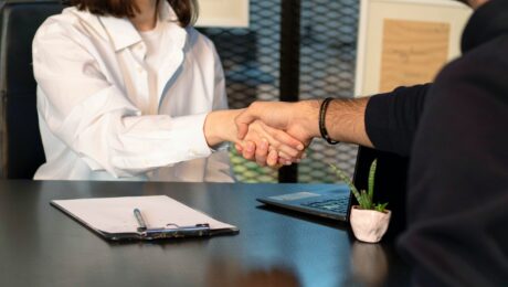 Two business leaders shaking hands across a table with a laptop and clipboard