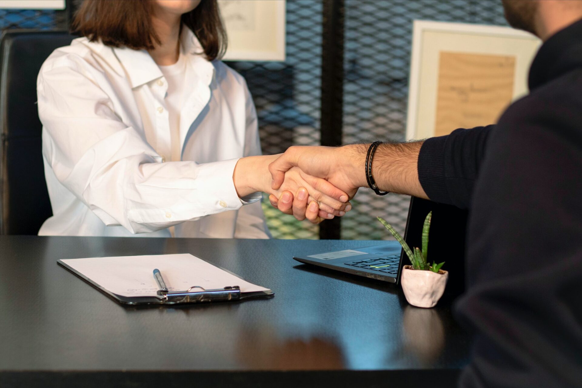 Two business leaders shaking hands across a table with a laptop and clipboard