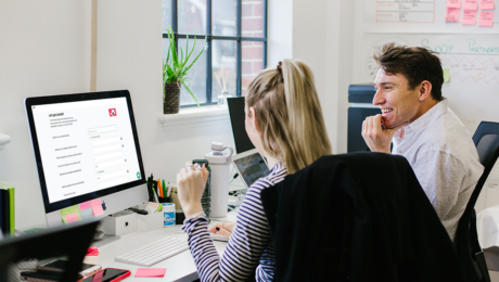 A man and a women looking at their laptop, while sat in front of a wall with sticky notes on