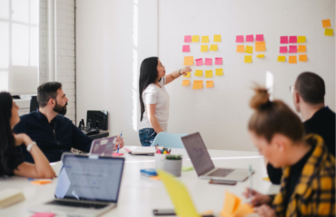 Women stood by wall of post it notes explaining to a group sat down what is on the notes