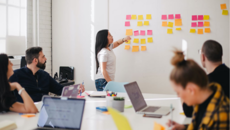 Women stood by wall of post it notes explaining to a group sat down what is on the notes