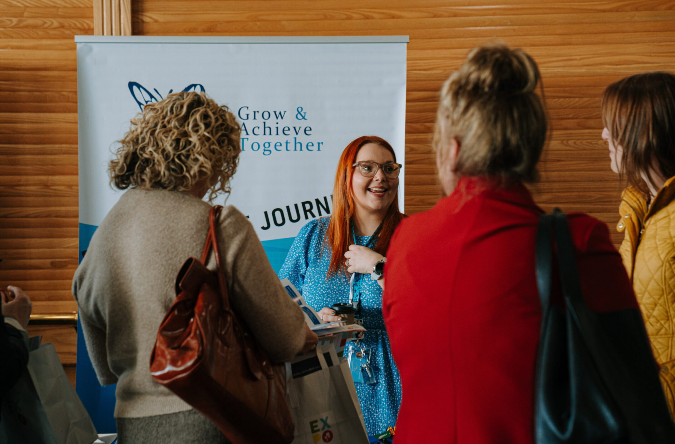 A woman with red hair, wearing glasses and a blue dress, smiling and interacting with attendees at a booth with a banner that reads 'Grow & Achieve Together' during a networking event.