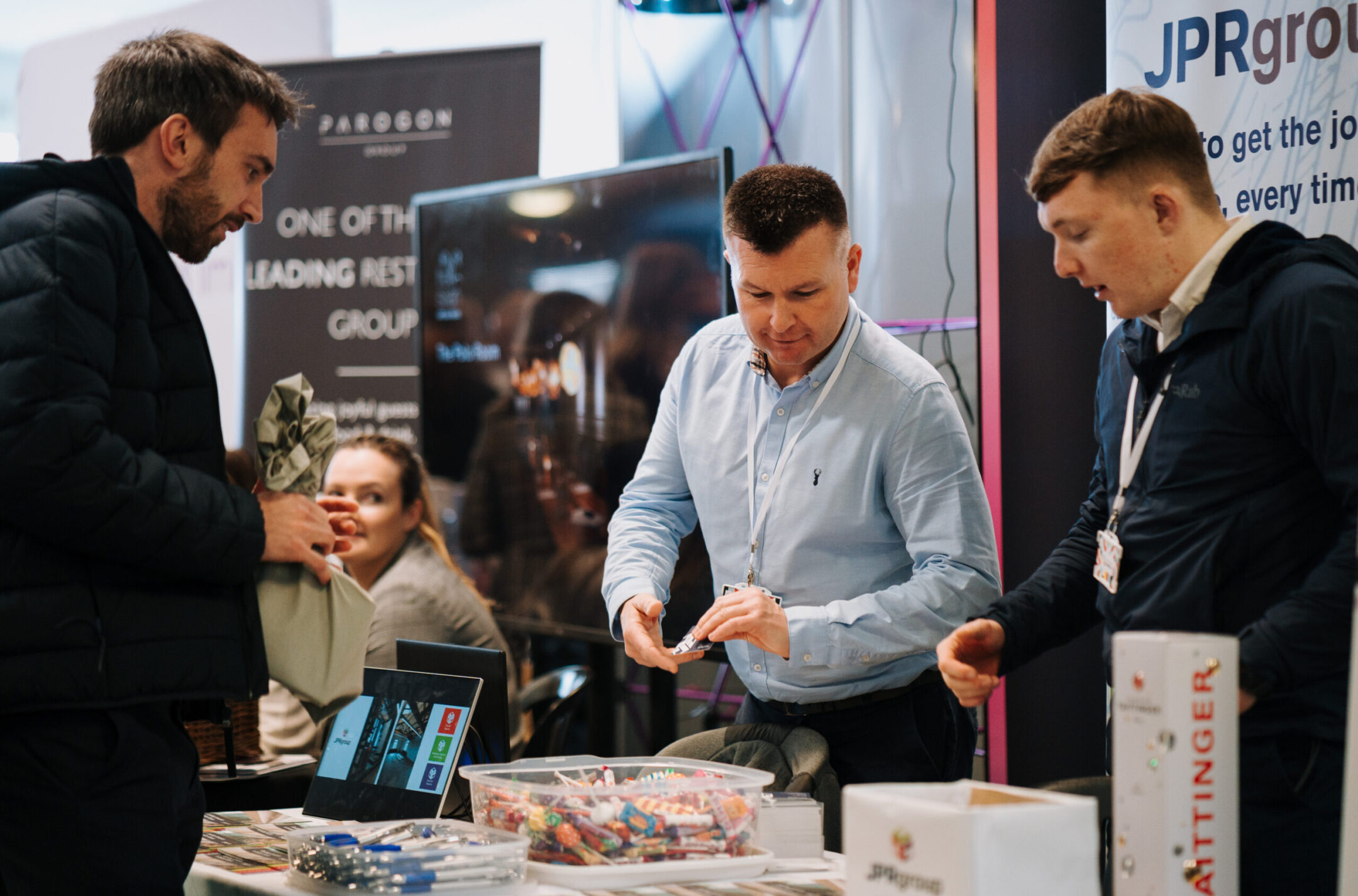 Two men engaging with a visitor at a booth during a networking event, with promotional materials, sweets, and a laptop on display, while a woman in the background observes the interaction.