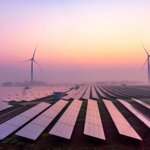 Picture of a solar panel farm with some wind turbines behind them.